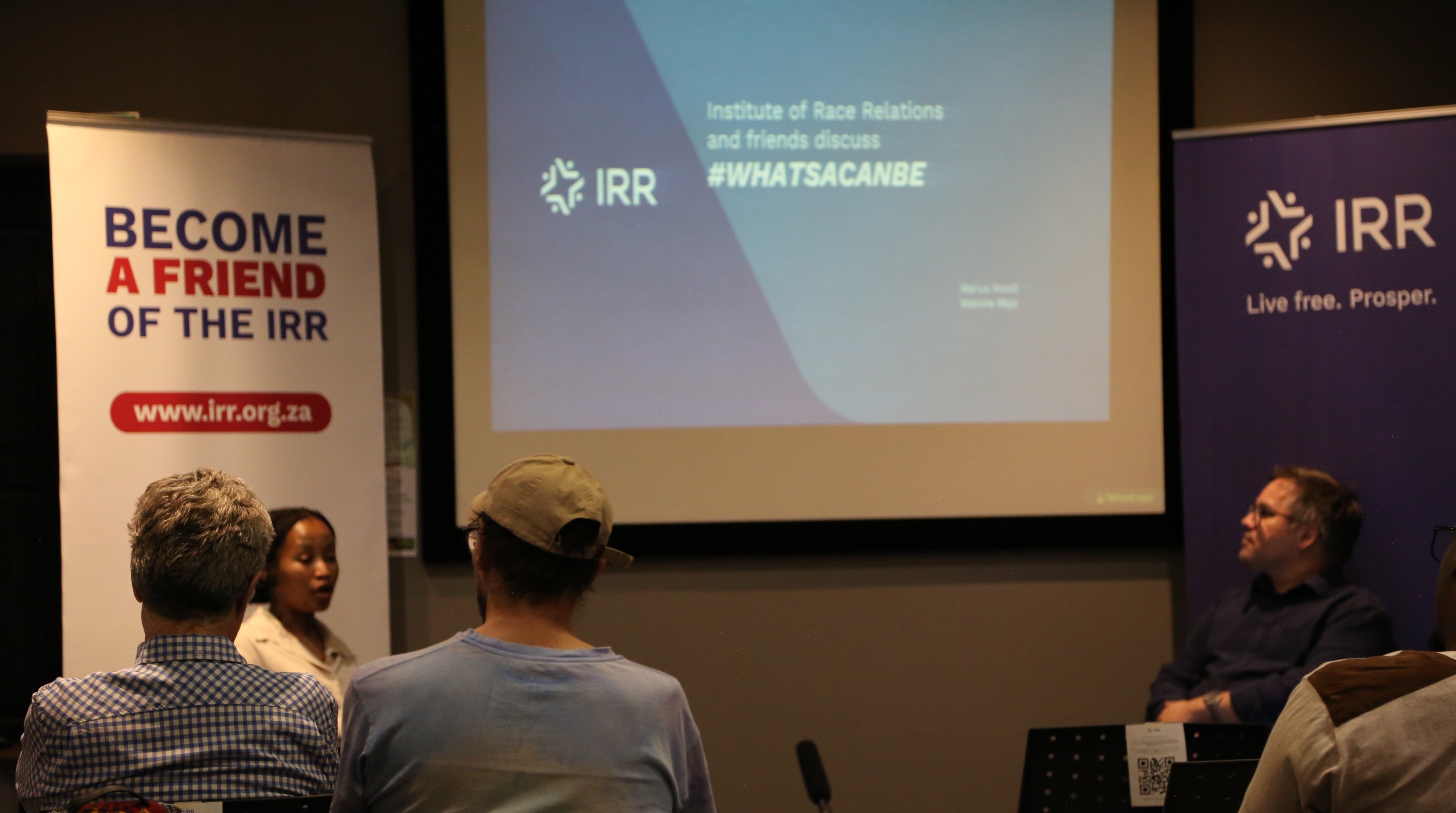 : A photograph of Marius and Makone studying a line graph on a slide being projected on a screen. There is a banner with the IRR logo in the background.